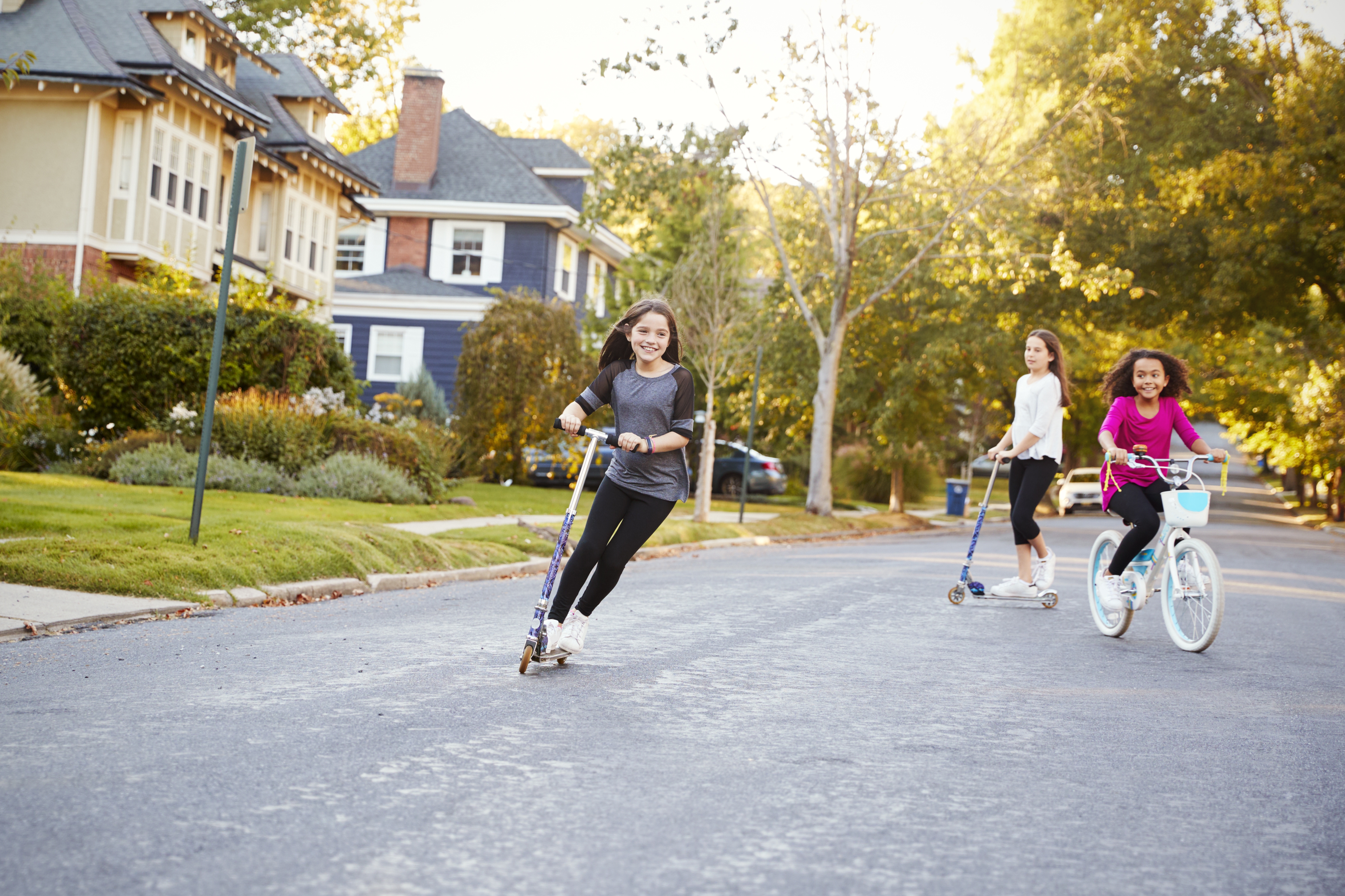 Three children riding scooters and a bike down a suburban street, enjoying a sunny day