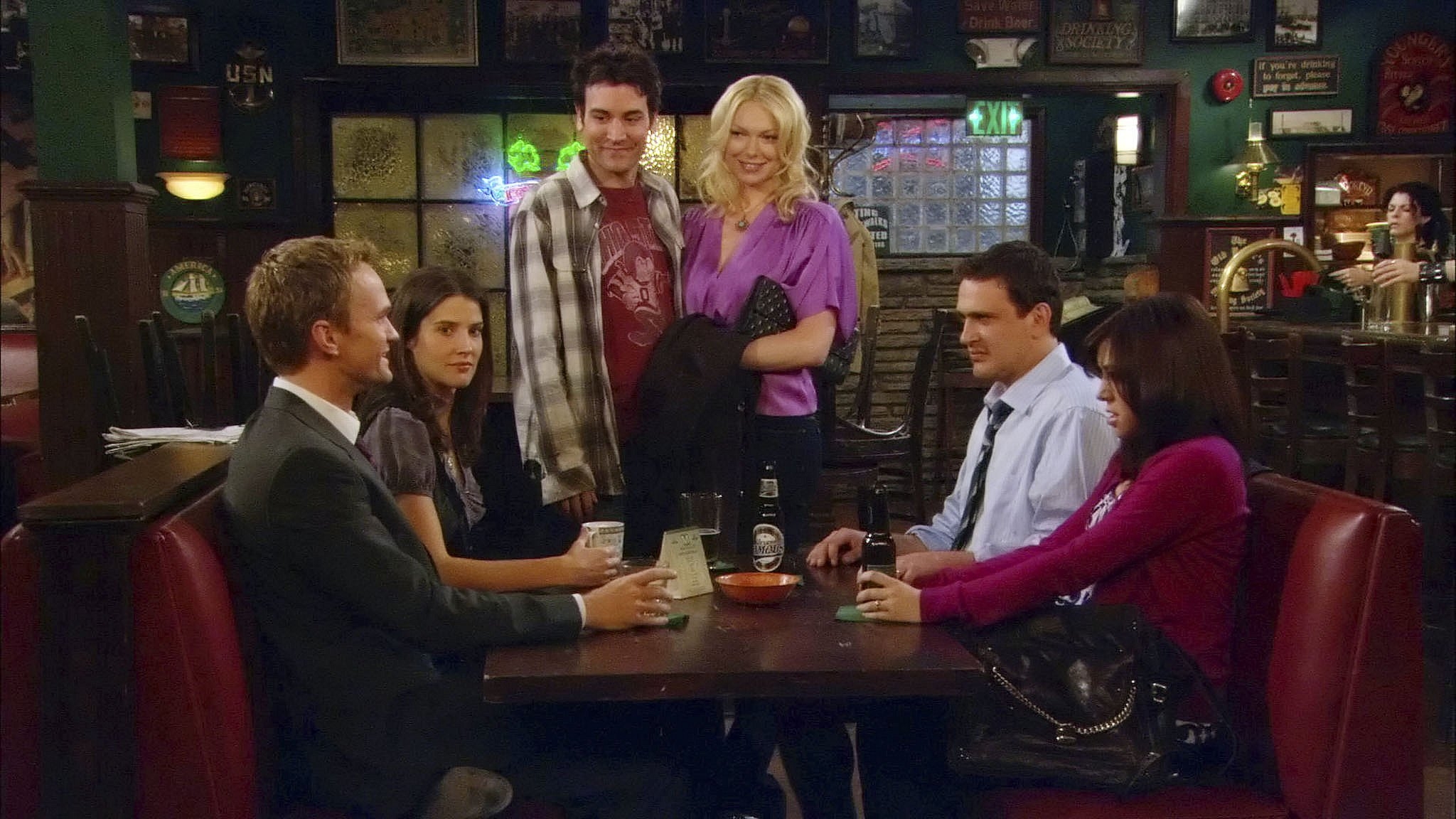 Group of six adults sitting and standing around a booth in a cozy pub setting, chatting and smiling casually