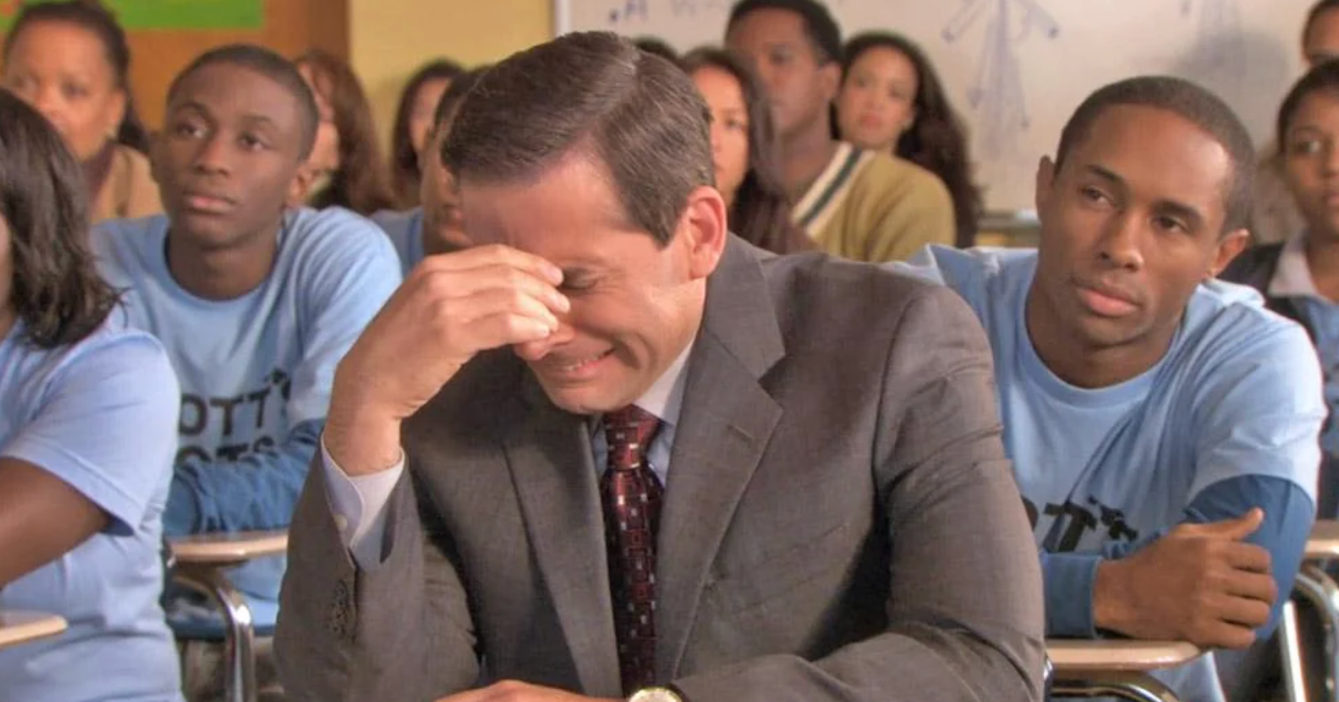 A man in a suit and tie sits in a classroom, appearing amused or frustrated, surrounded by students in casual shirts