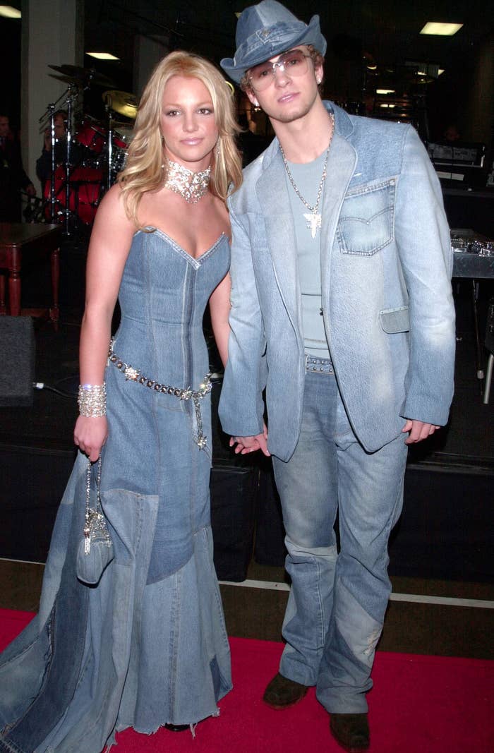 Two people in matching denim outfits on a red carpet. One is wearing a denim gown, the other a denim suit with a cowboy hat
