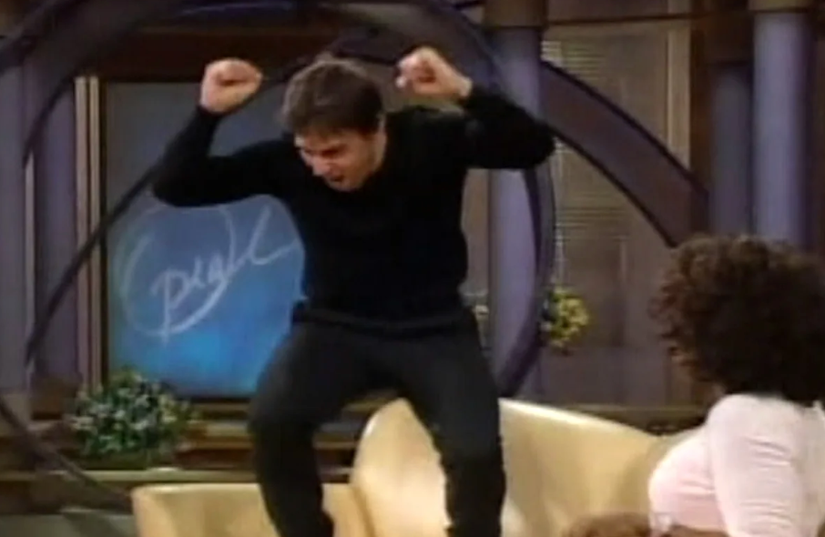 A person energetically jumping on a couch during a talk show while the host looks on with interest