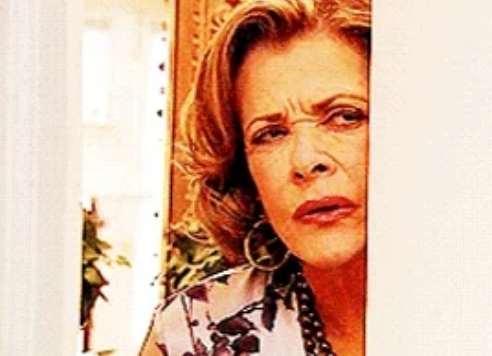 Person peering from behind a wall with a curious expression, wearing a floral-patterned top and hoop earrings