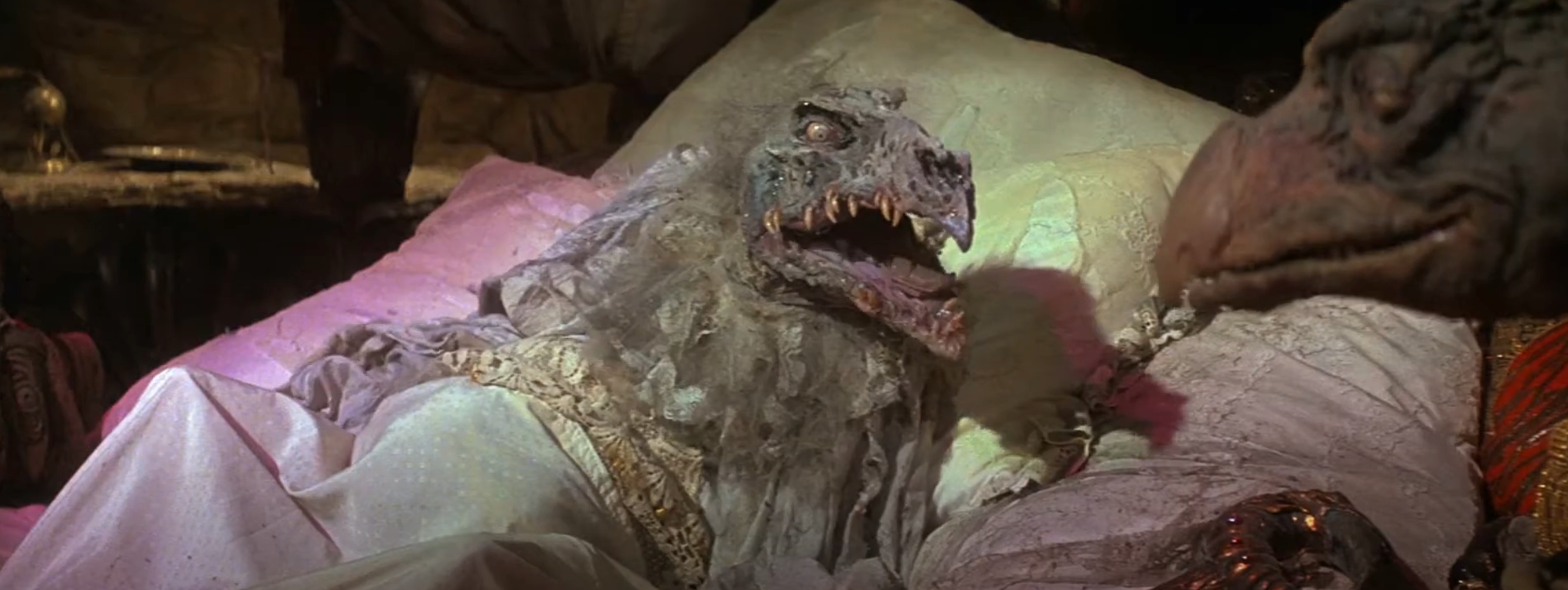Creature in elaborate costume resting on a bed, interacting with another similar character inside a dimly lit room