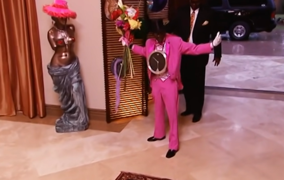 Person in oversized suit with clock necklace and wide-brimmed hat, standing indoors