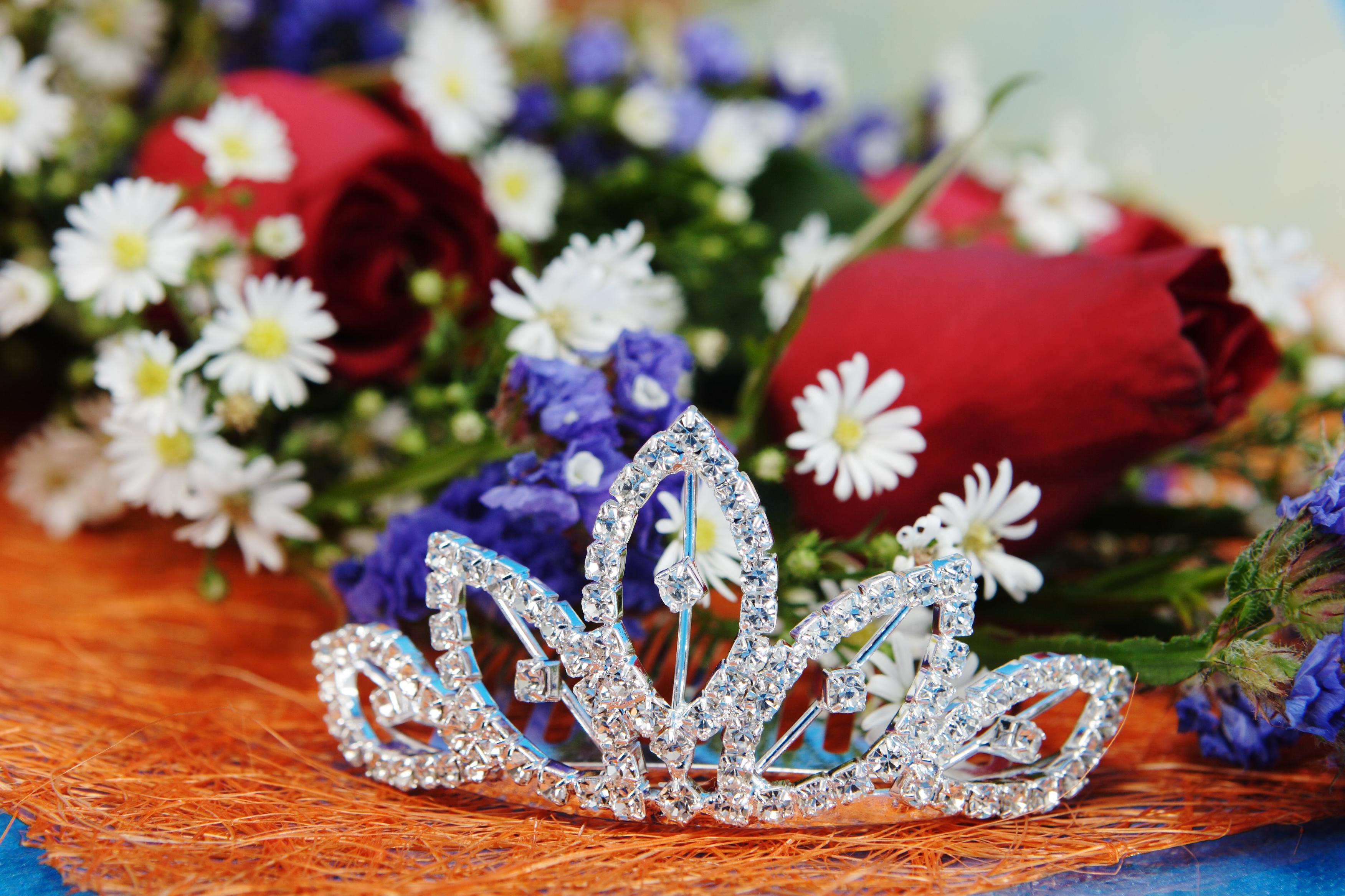 A sparkling tiara is set in front of an arrangement of roses and white daisies.