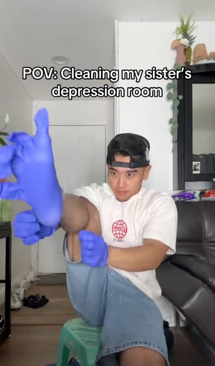 Person in casual clothes sits on a stool, wearing gloves, preparing to clean a room. Text: "POV: Cleaning my sister’s depression room."
