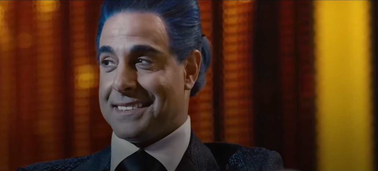 Man with slicked-back hair smiles in an interview setting, wearing a formal suit and tie. A dramatic background adds to the scene's intensity