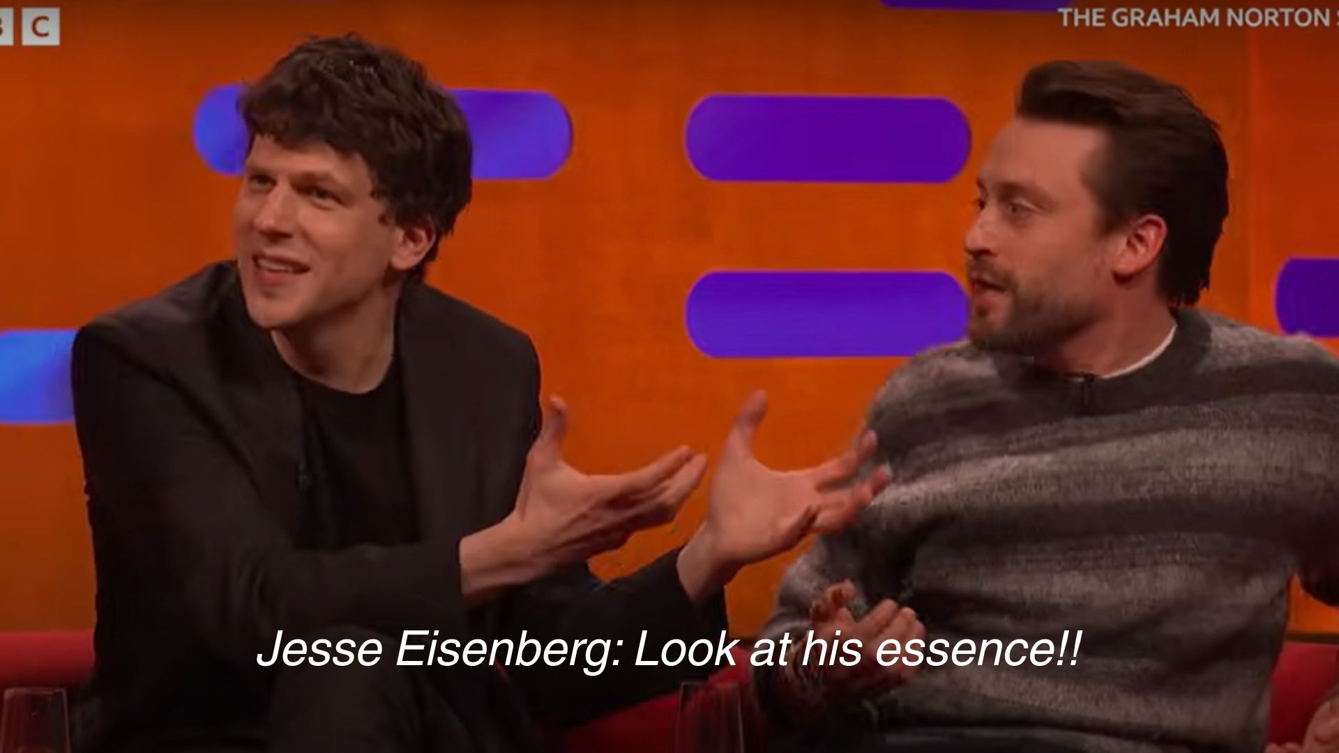 Two men sit on a talk show set, one animatedly gesturing while the other listens. Subtitles read: "Jesse Eisenberg: Look at his essence!!"