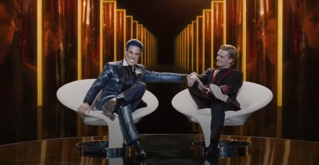 Two people seated on stylish chairs, smiling and shaking hands, set against a backdrop of illuminated columns