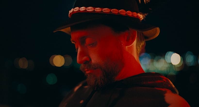 A person wearing a hat decorated with shells is looking down against a blurred night background with distant lights