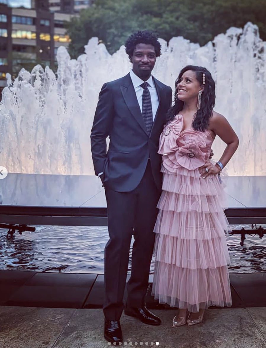 Two individuals stand in front of a fountain. One wears a suit, the other a tiered dress with floral detail, both smiling for the camera