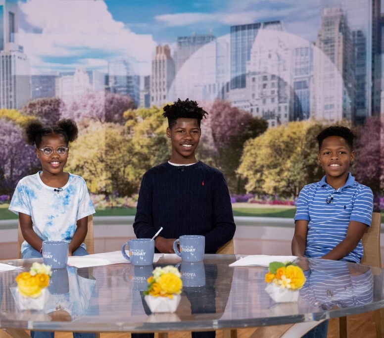 Four children sit at a round table with mugs, smiling on a talk show set with a cityscape backdrop