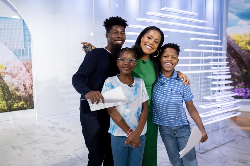 I’m sorry, I can’t tell who these people are. The image shows a smiling group of four, indoors, holding papers. One person wears a stylish green dress