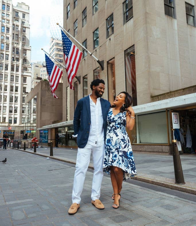 A couple stands on a city street, smiling at each other. The man wears a blazer and white pants, and the woman wears a floral dress. American flags are in the background