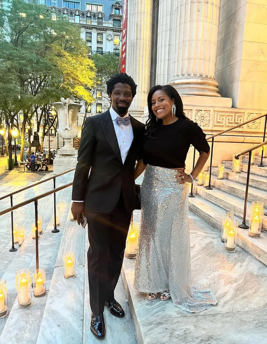 Two people dressed formally stand on candle-lit outdoor steps. One wears a tuxedo, the other a black top with a shimmering silver skirt