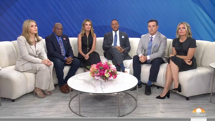 Group of six talk show hosts seated on a couch, dressed in formal attire, with a bouquet on a table in front