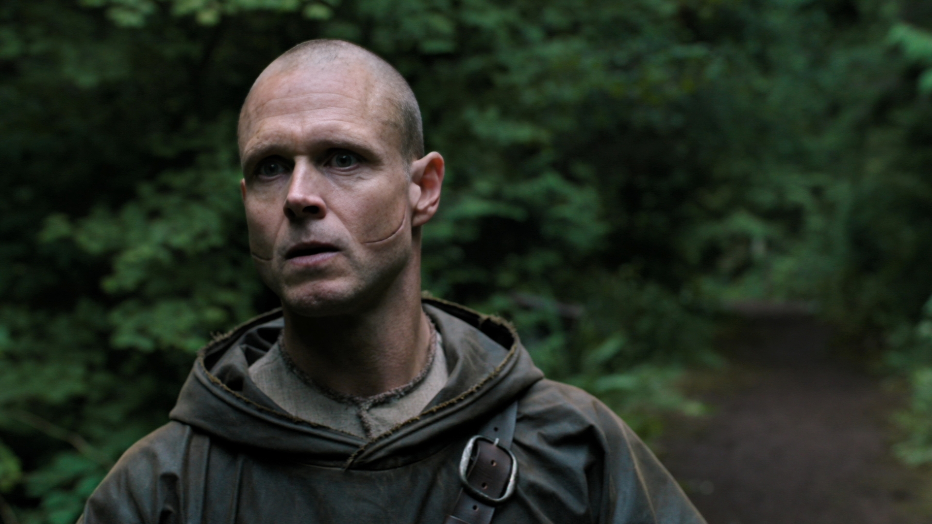 Persona calva en un bosque con expresión seria, lleva un abrigo con capucha. Fondo de follaje denso