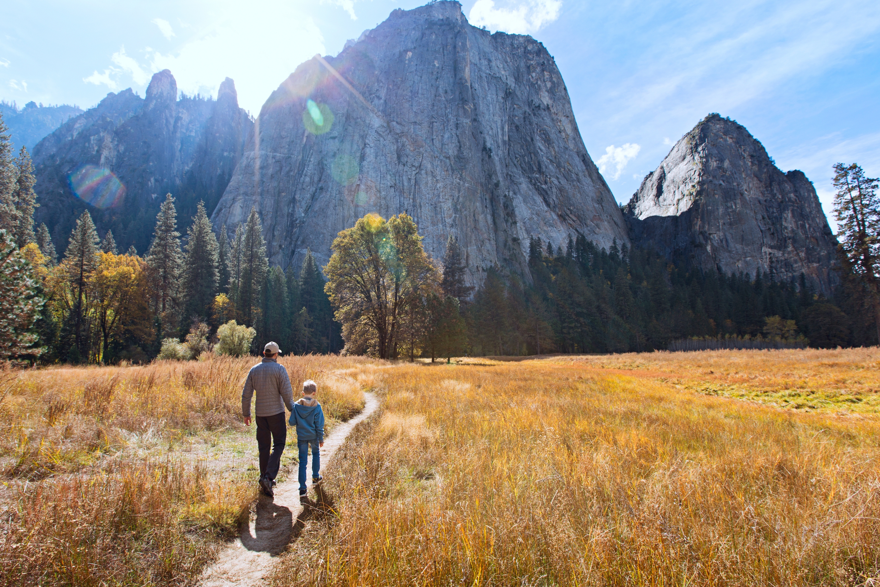 Two people walk on a path through a meadow toward towering mountains under a clear sky