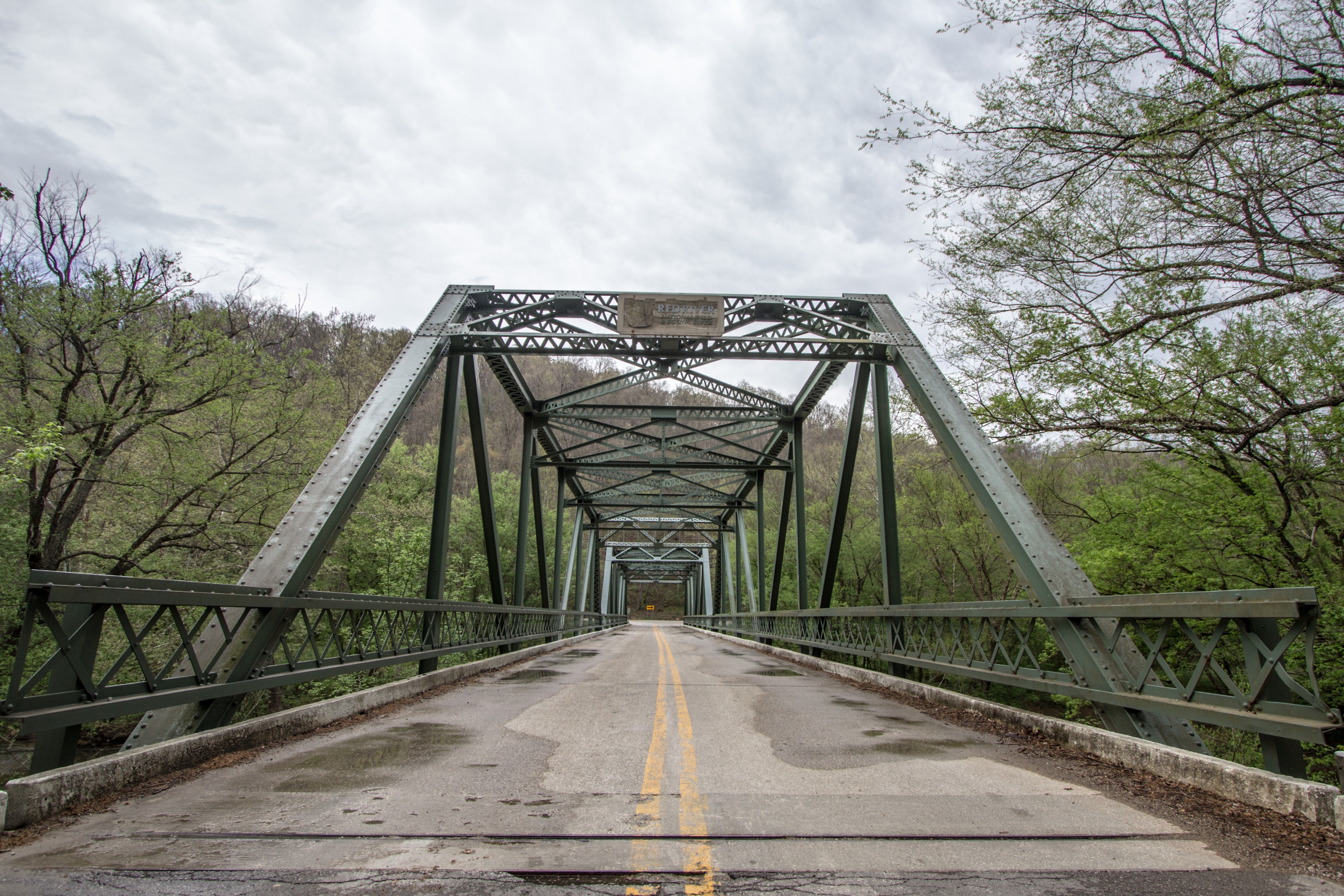 Steel truss bridge with lattice railings spans a river, framed by trees. Overcast sky above