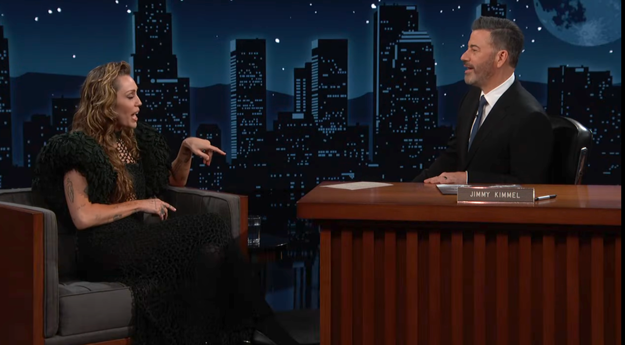 Talk show host interviews a guest wearing an elegant, textured dress on a nighttime set with a cityscape backdrop