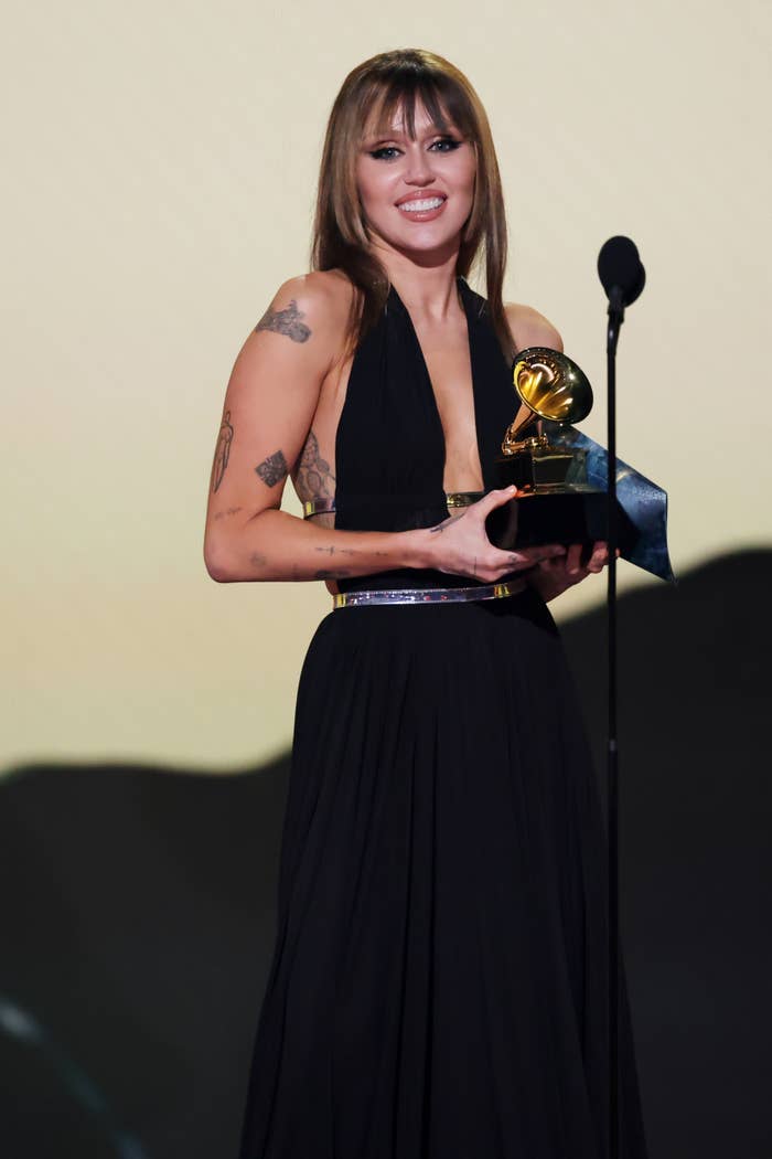 Person in a halter gown smiling, holding a Grammy award, standing at a microphone on stage