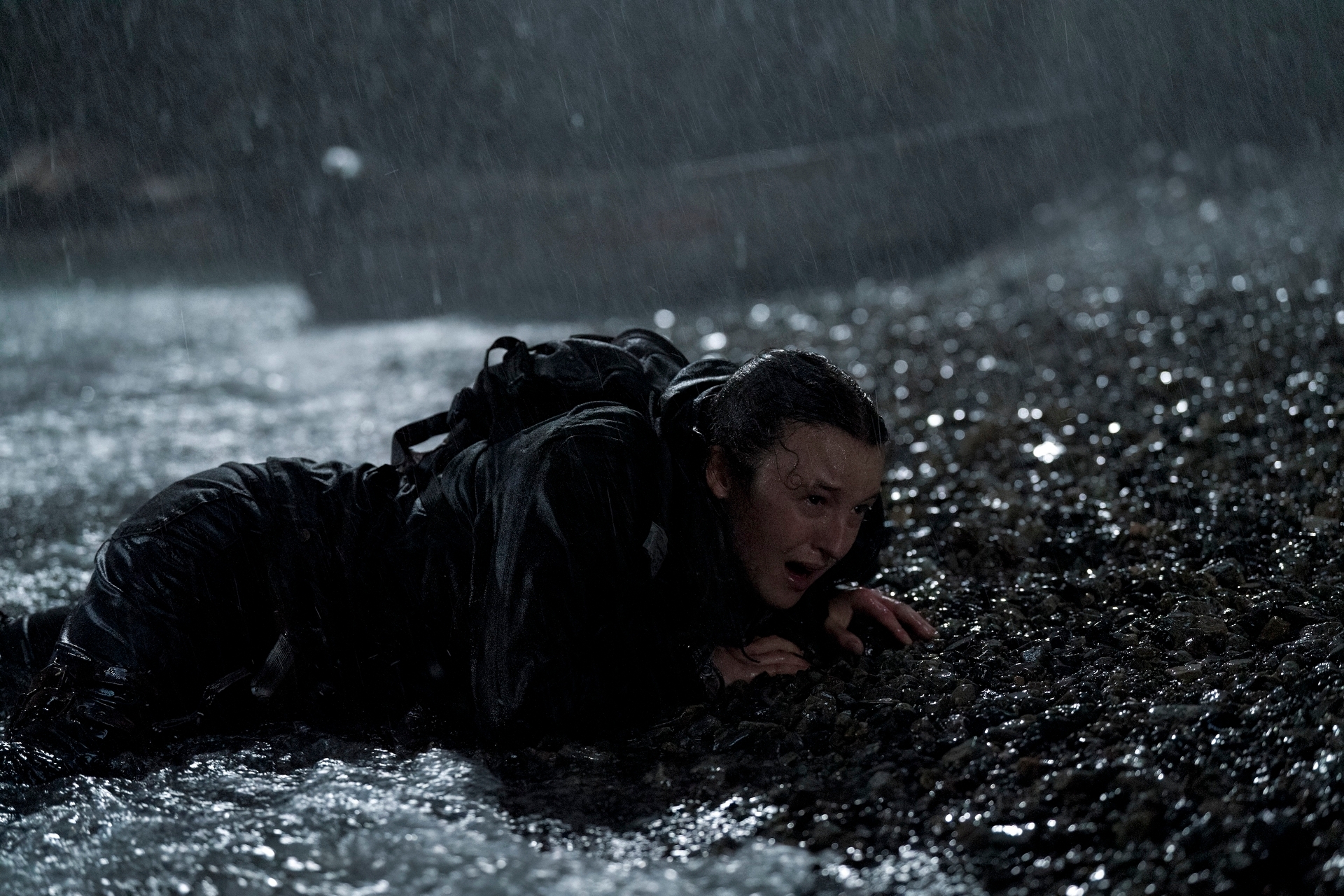 Persona en la orilla bajo la lluvia, con ropa mojada y expresión de esfuerzo, arrastrándose en un escenario oscuro y pedregoso