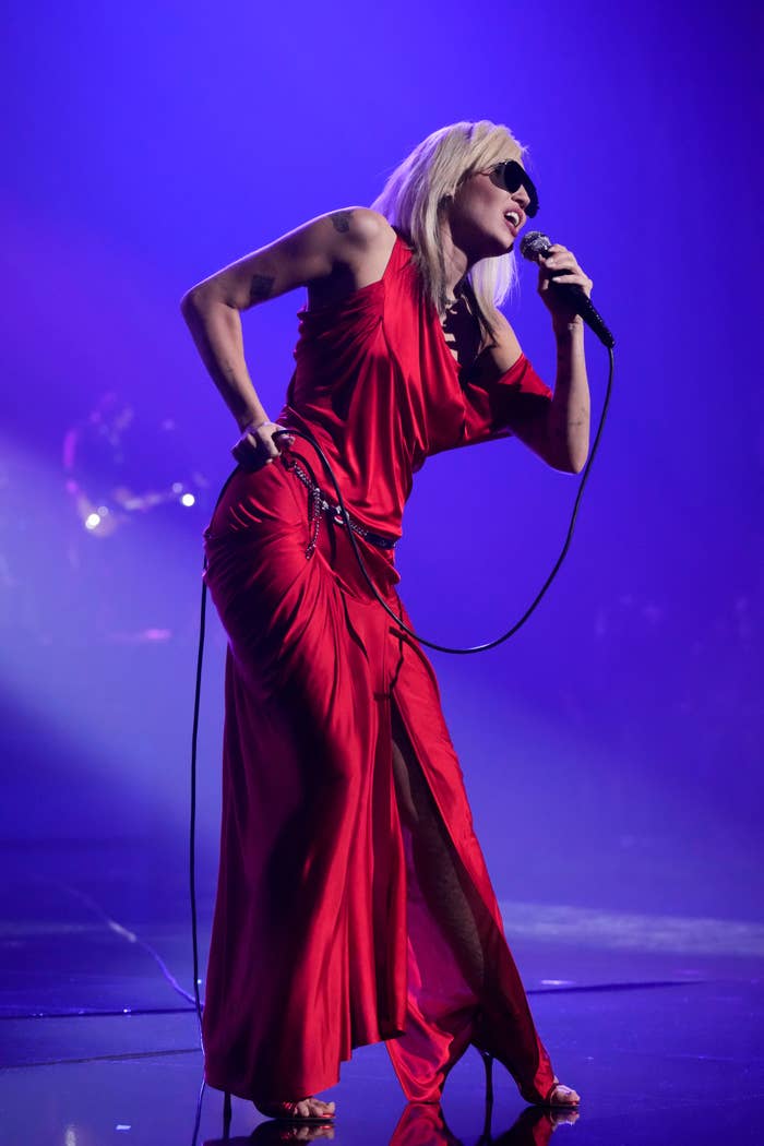 Person performing on stage in a red satin gown with side slit, wearing sunglasses and holding a microphone passionately