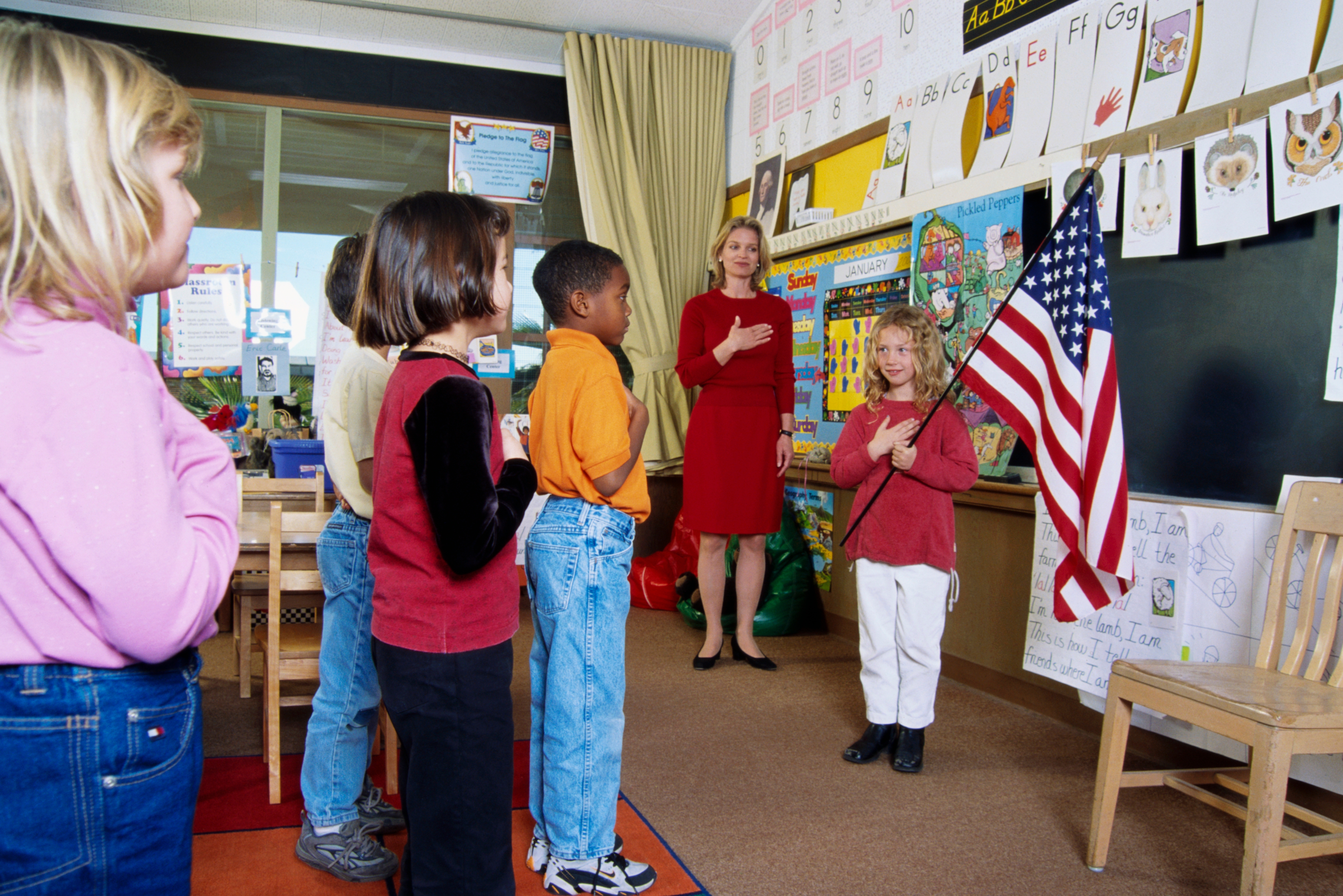 Children stand with their hands over their hearts by a teacher in a classroom, reciting the pledge of allegiance near a U.S. flag