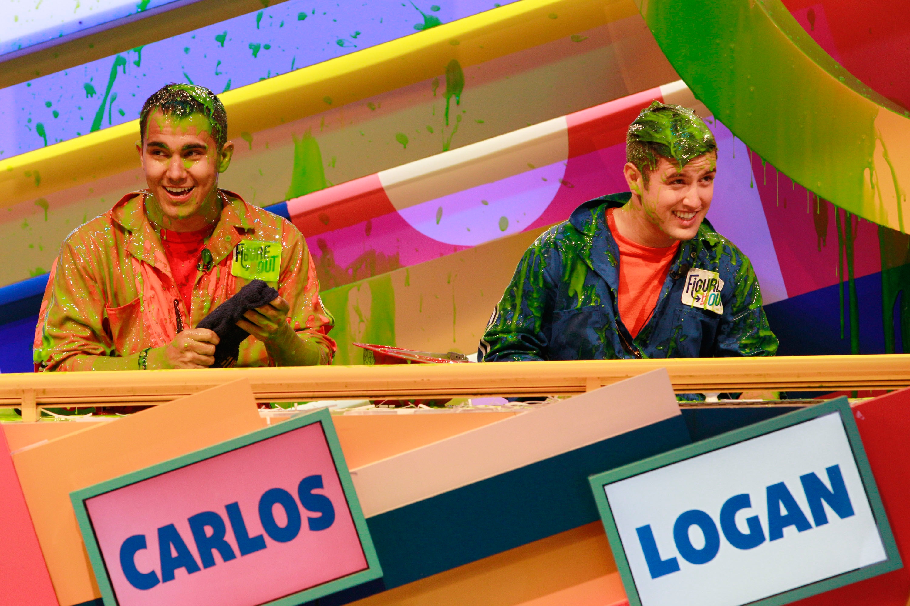 Two people in slimy game show attire are covered in green slime. They stand cheerfully behind a railing with "Carlos" and "Logan" name tags