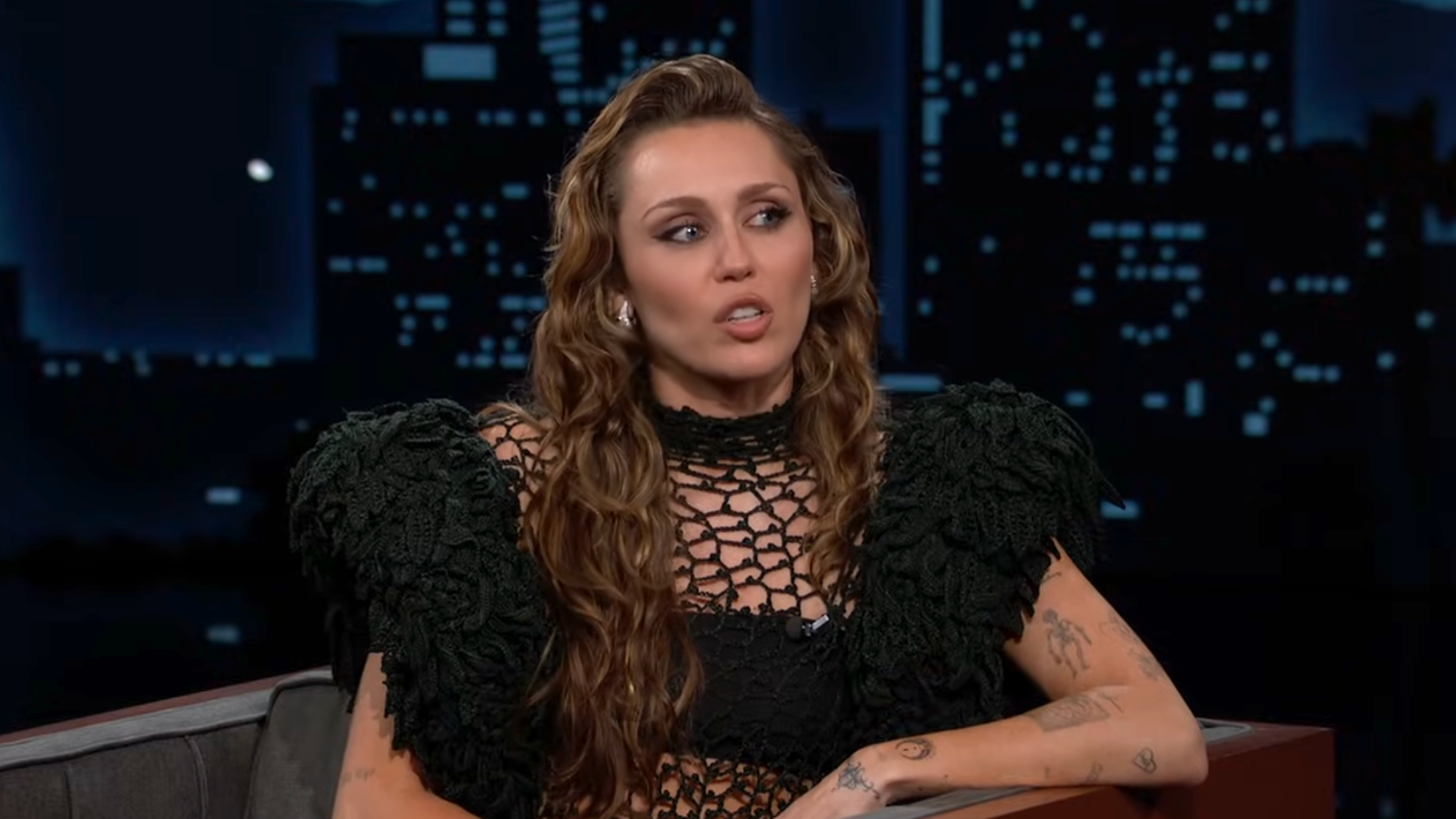 A person with long wavy hair sits on a talk show set, wearing a unique crocheted top with puffed shoulders