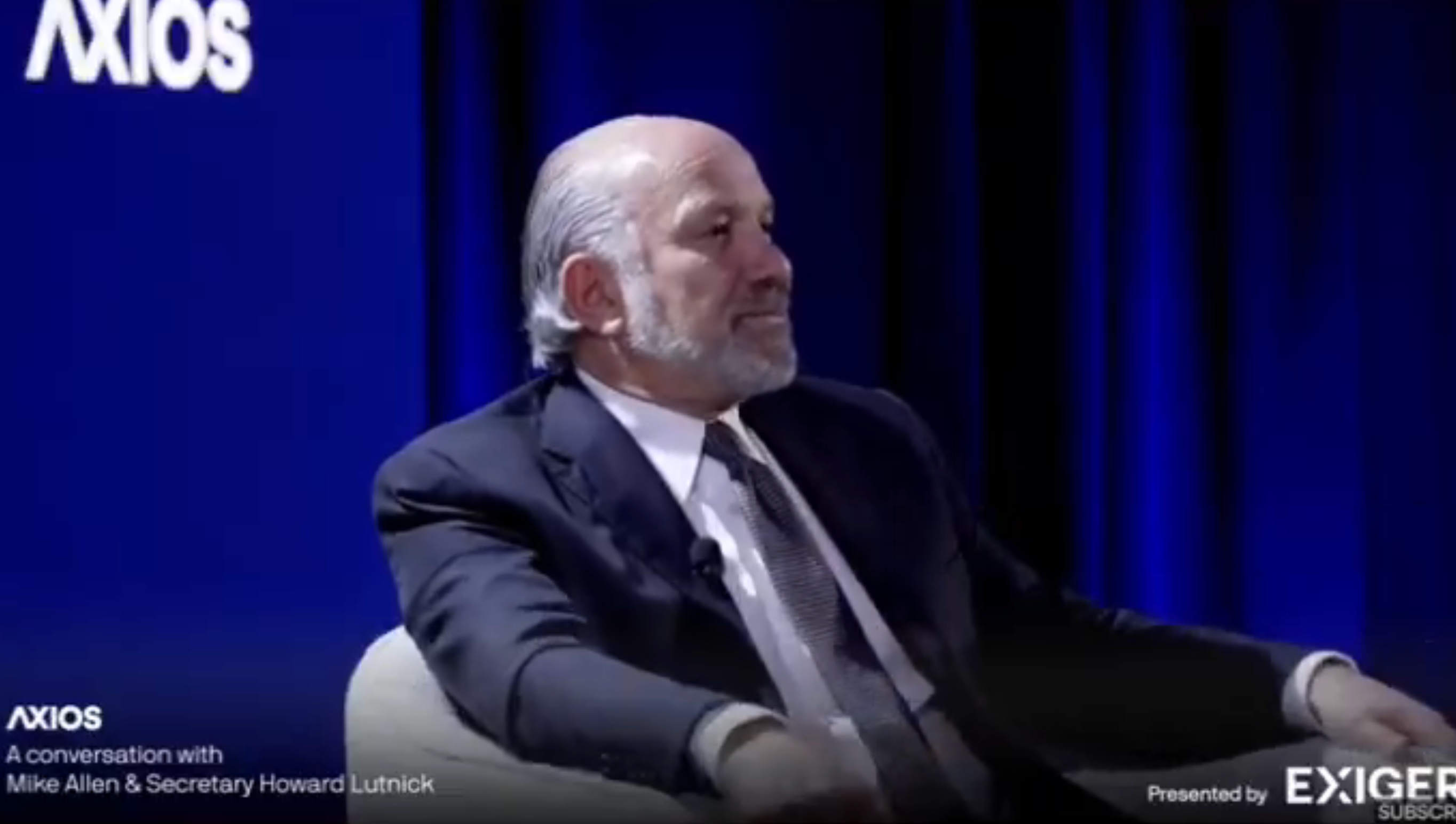 A man in a suit sits on stage during an Axios conversation event, engaging in a discussion with an unseen person