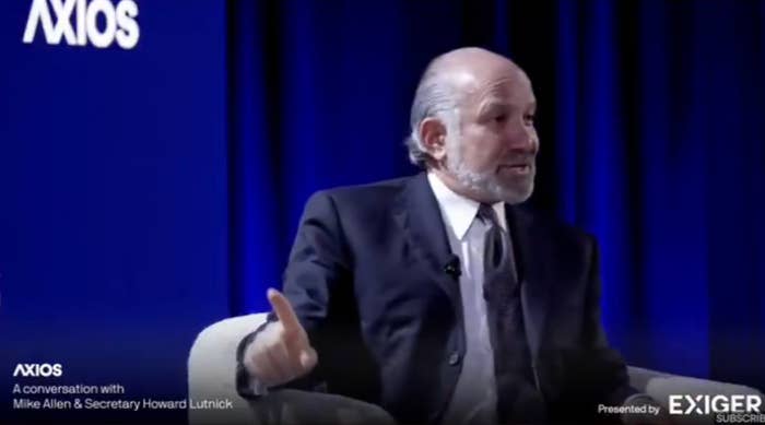 Person in a suit speaking onstage in a panel discussion setting, blue backdrop. Text reads: &quot;AXIOS: A conversation with Mike Allen &amp;amp; Secretary Howard Lutnick.&quot;
