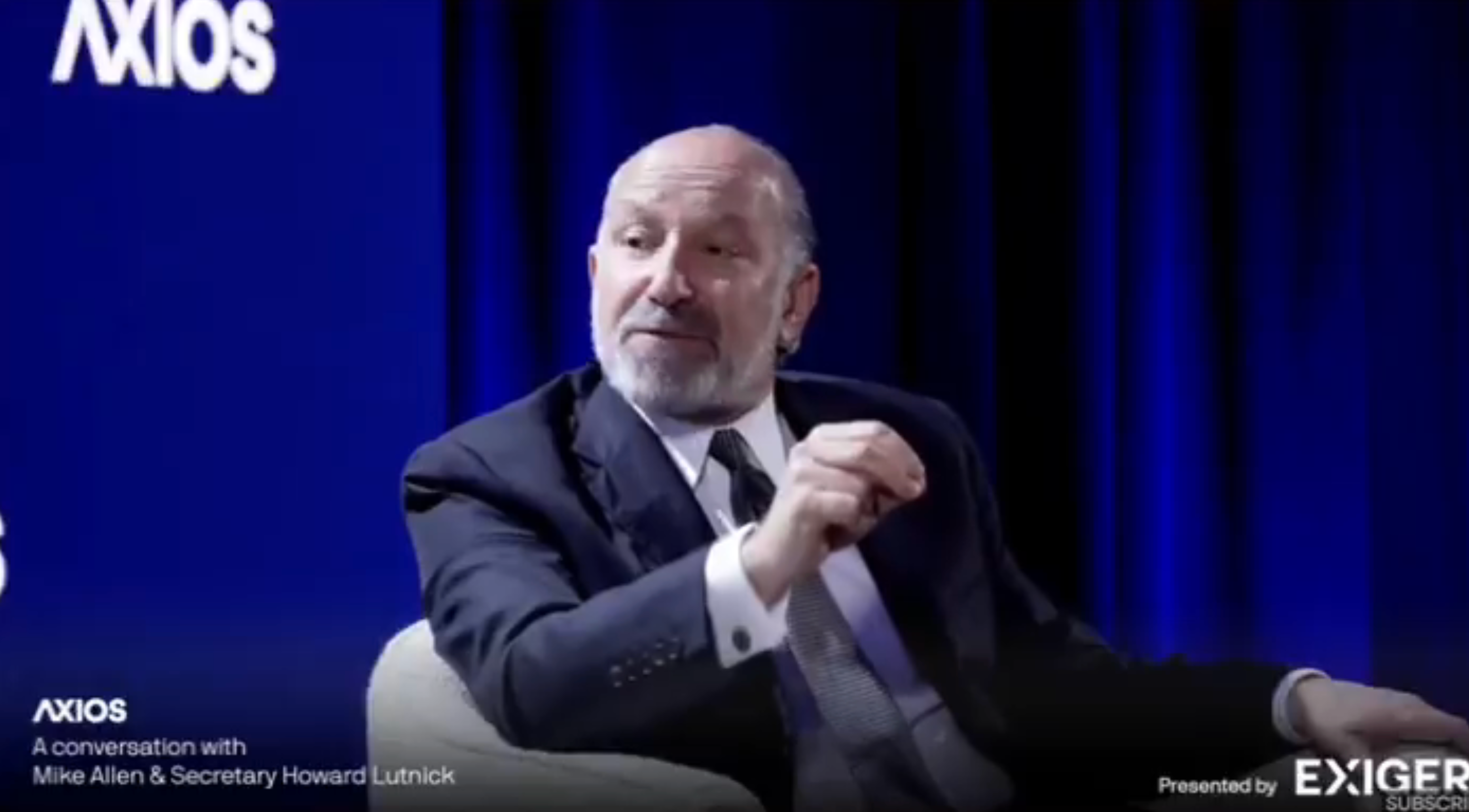 A man in a suit speaks during an Axios conversation event. A banner with the text &quot;Axios&quot; is in the background