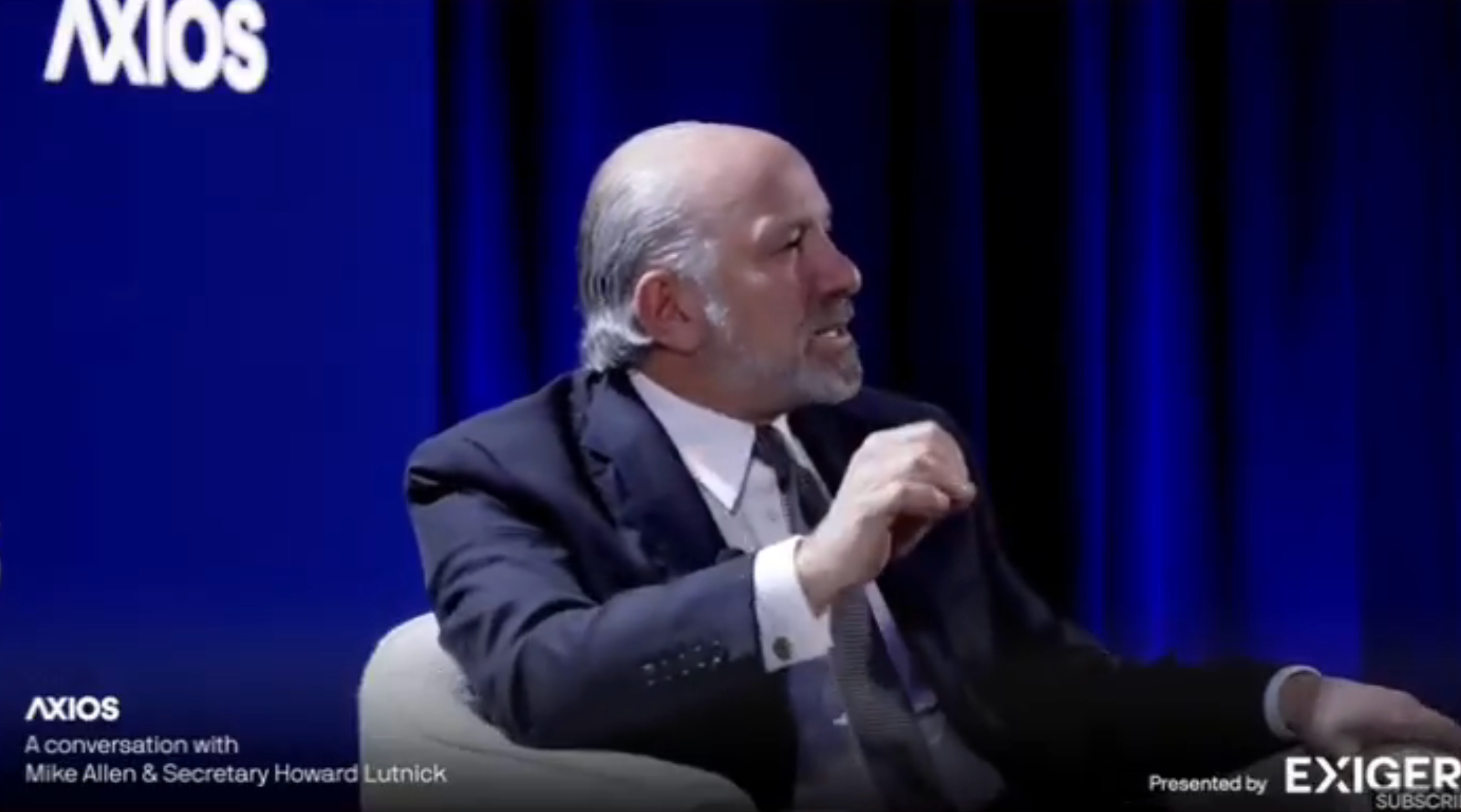 A man in a suit sits on stage during an Axios event, engaged in conversation