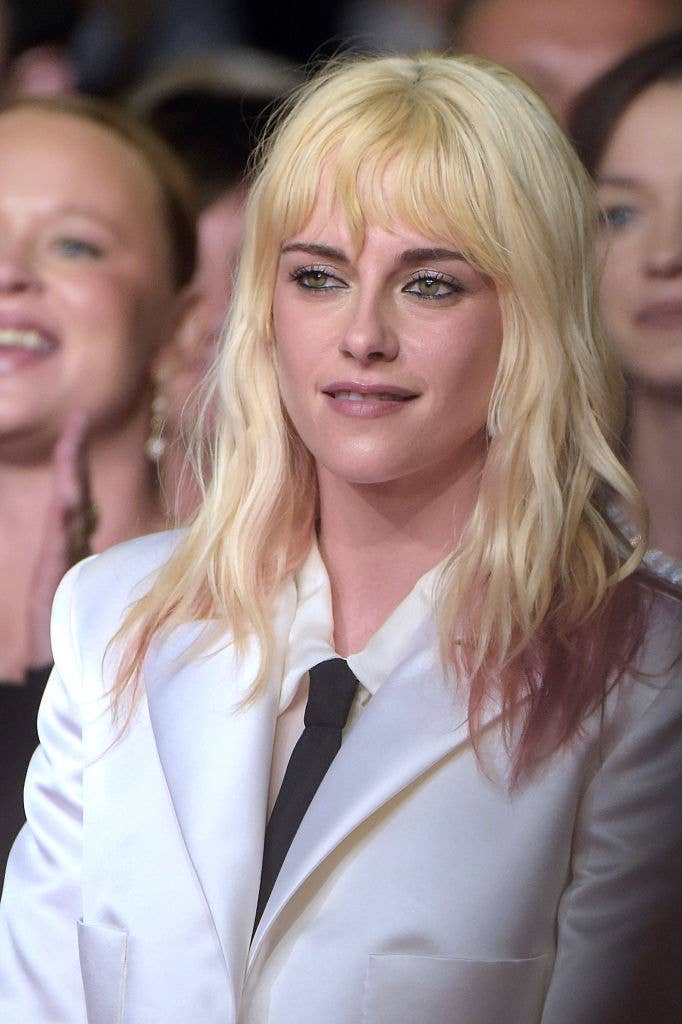 A person with shoulder-length hair wears a white suit with a black tie at a public event, surrounded by others