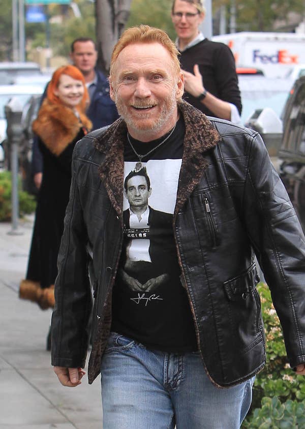 Man in a leather jacket with faux fur trim, graphic tee, and jeans, smiling while walking on a city sidewalk
