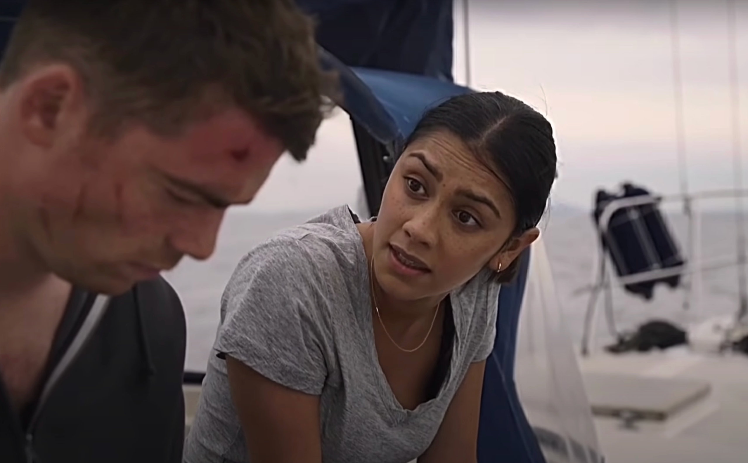 A woman and man, both looking concerned, sit on a boat. The man has visible scratches on his face. Scene suggests a tense moment