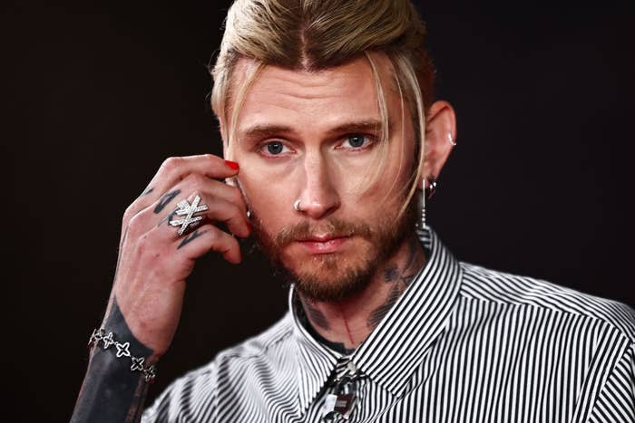 Person with tattoos and facial piercings, wearing a striped shirt, touching their ear