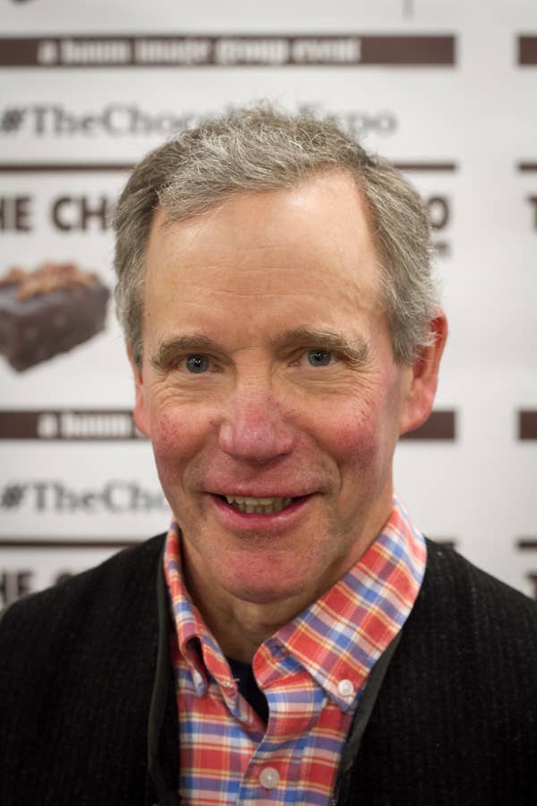 They are at an event with a checkered shirt under a sweater, in front of a backdrop with text and a chocolate image