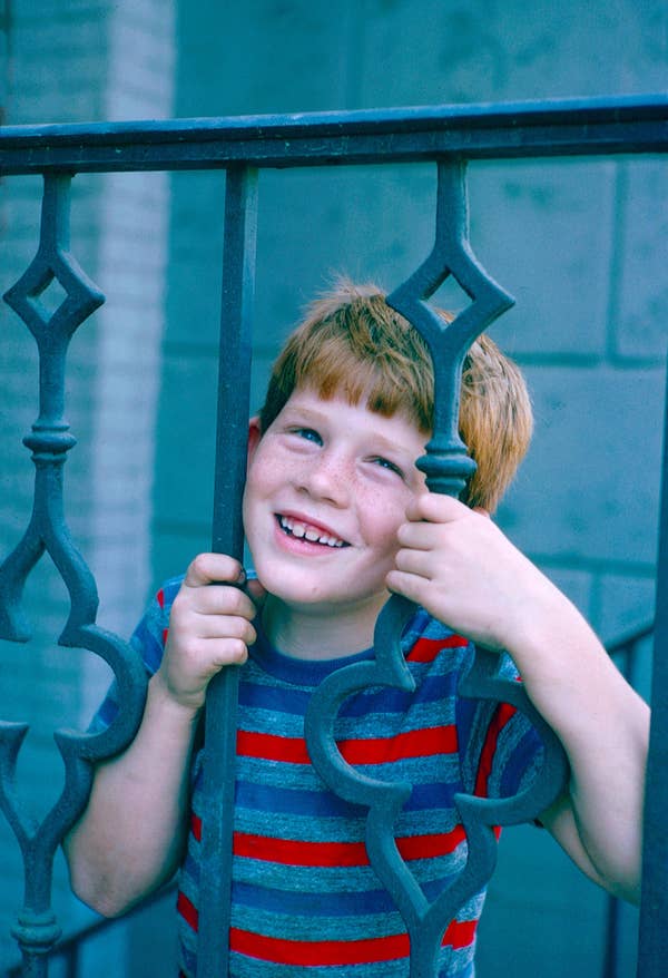 A smiling child with short hair playfully peers through a decorative metal railing, wearing a striped shirt
