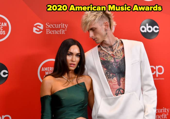 Two celebrities on a red carpet; one in a green off-shoulder dress, the other in a white suit with tattoos, looking at each other