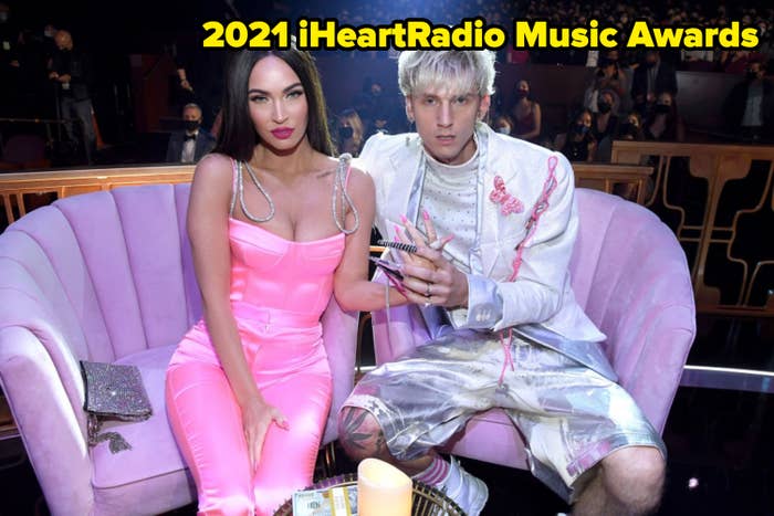 Two celebrities seated on a couch, one in a fitted jumpsuit and the other in a contemporary suit with shorts, holding an award. Audience visible behind