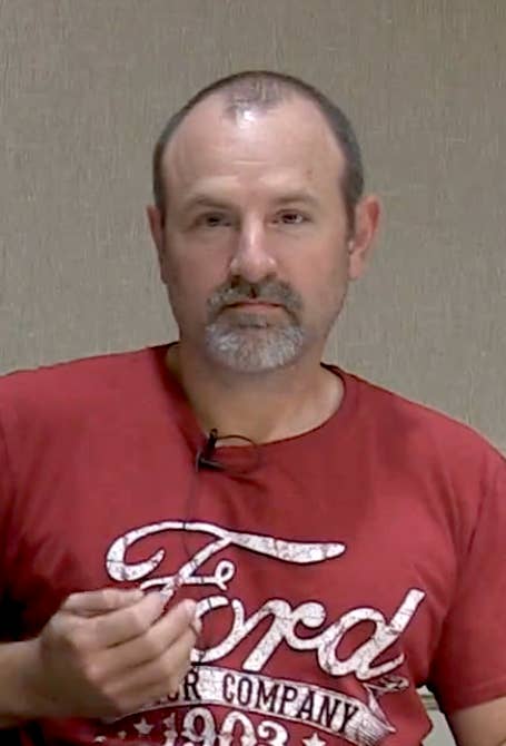 Man seated at a table, wearing a casual t-shirt with a "Ford Motor Company" design, speaking into a microphone