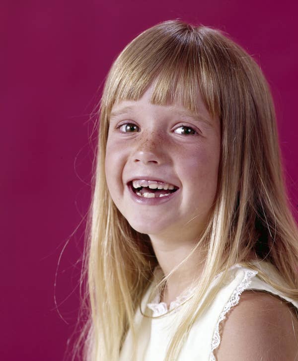Smiling young girl with long blonde hair and bangs, wearing a lace-trimmed dress, posed against a solid background