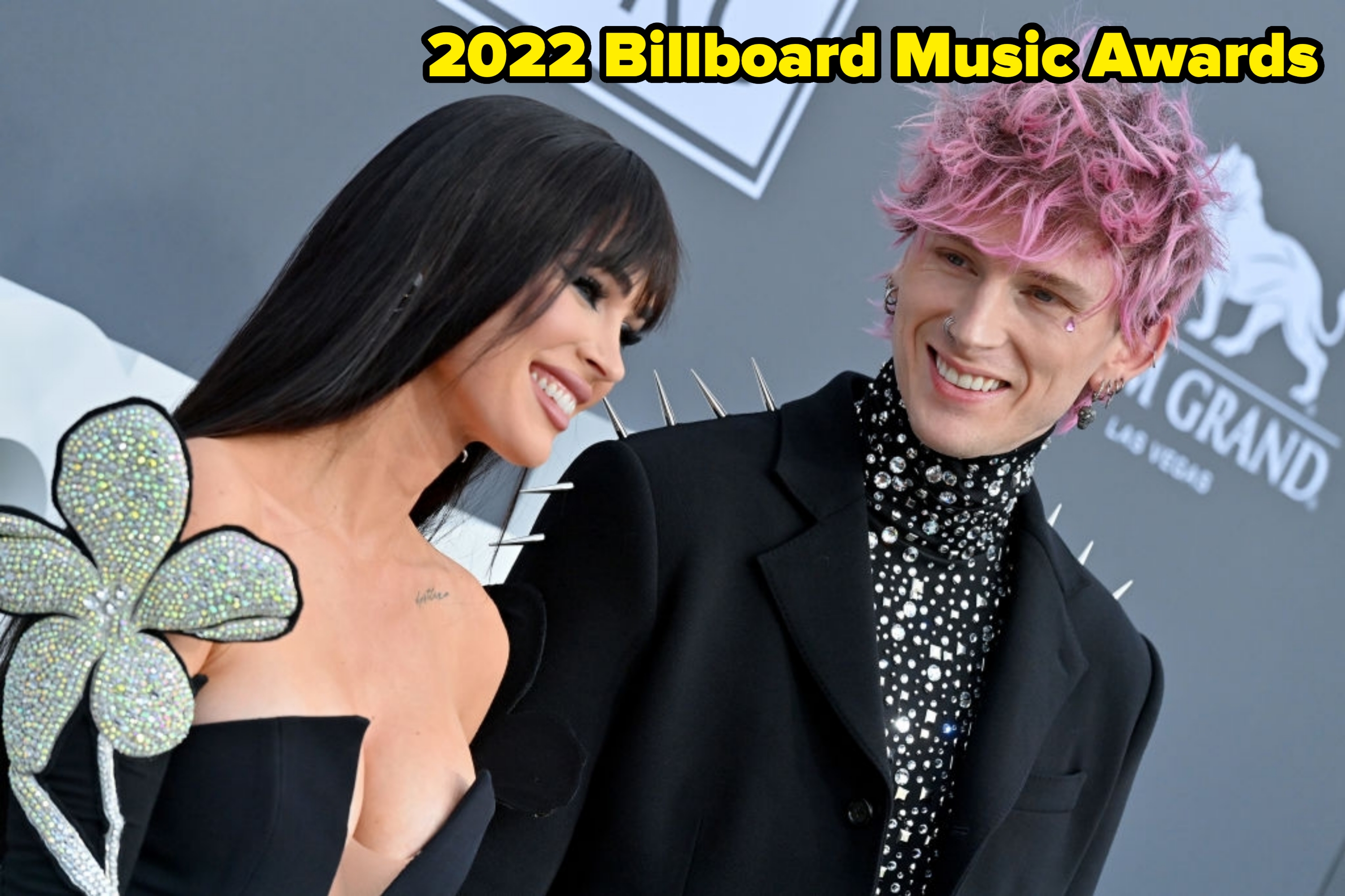 Two people smiling on the red carpet. One wears a floral-themed black dress; the other, a black suit with spikes and a spotted shirt