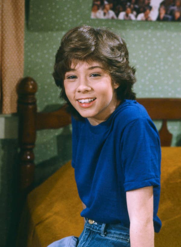 A young person with medium-length hair sits on a bed, smiling. They wear a casual t-shirt and jeans