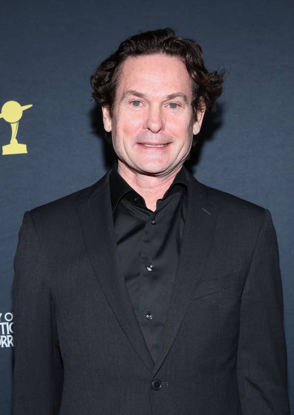 Person in a tailored suit attends the Saturn Awards, standing in front of a backdrop with event logos