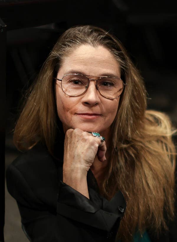 Person sitting in a stadium setting, wearing a black blazer over a teal top, with glasses and long hair. They rest their chin on their hand