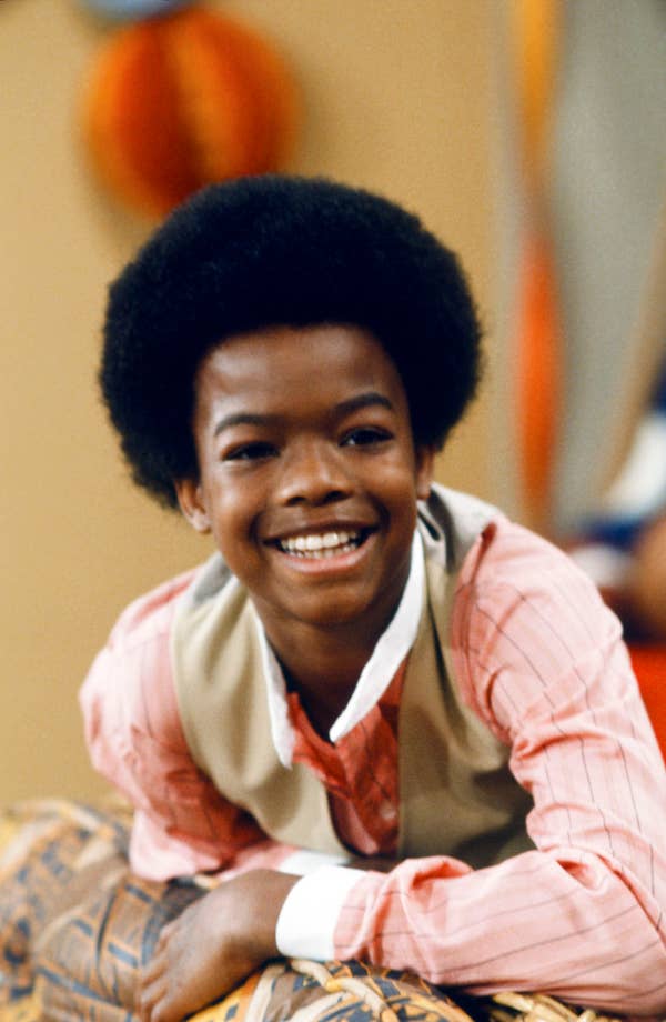 A young person with an afro hairstyle smiles, wearing a pink shirt and beige vest, leaning on a patterned surface