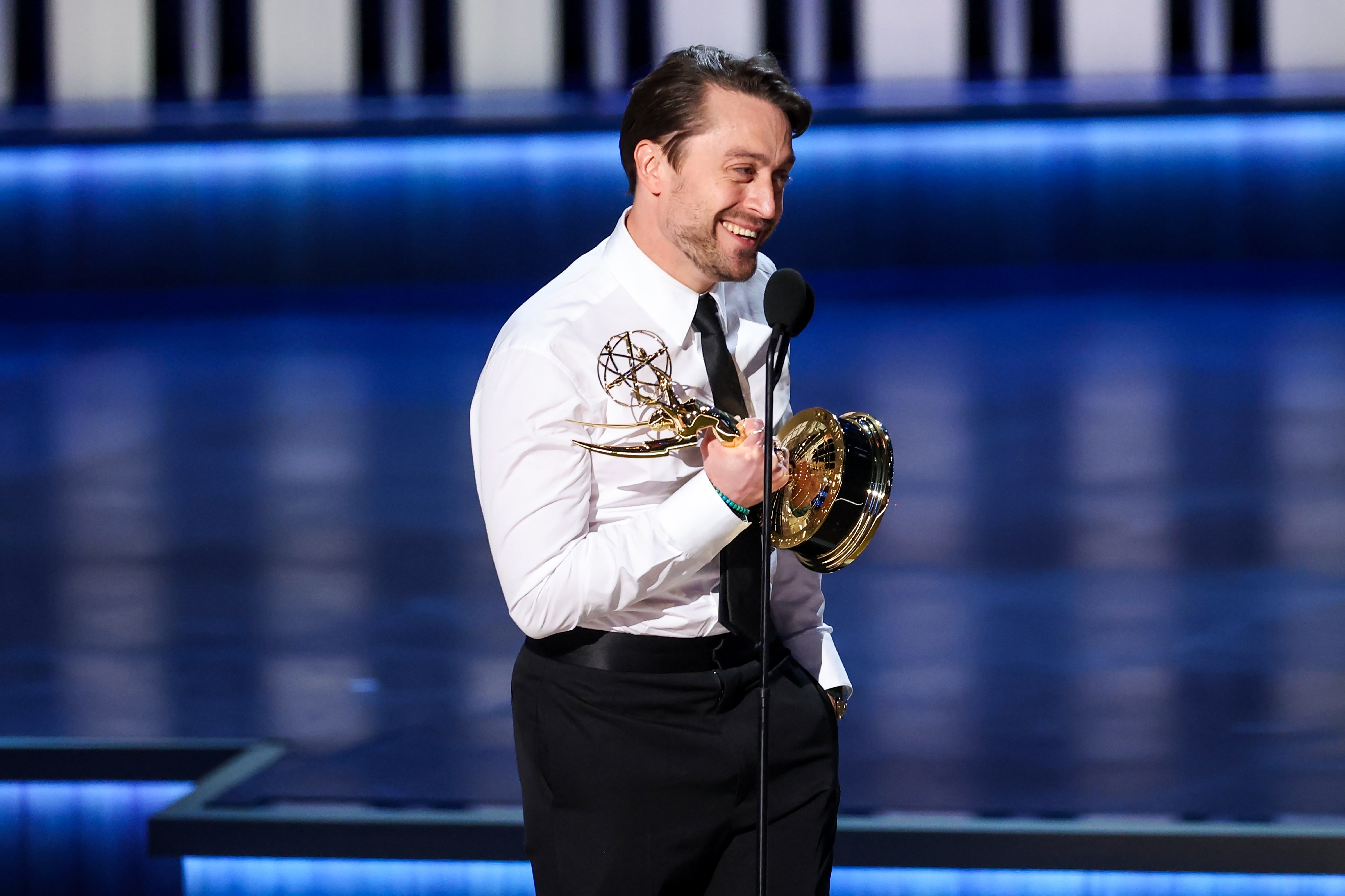 Man in formal attire holding Emmy award, smiling at microphone on stage